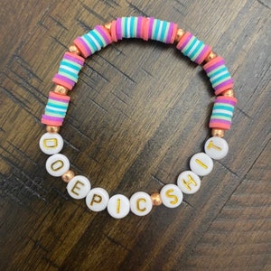 May include: A colorful beaded bracelet with pink, blue, and white striped beads and white letter beads spelling out "DO EPIC SHIT".