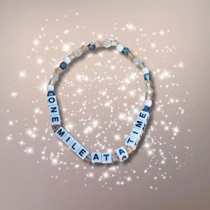 May include: A white beaded bracelet with gold and blue accents. The beads spell out the phrase "One Mile At A Time".
