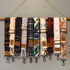 Pode incluir: Uma coleção de alças de bolsa estampadas penduradas em uma vara de madeira. As alças apresentam vários designs, incluindo camuflagem, estrelas, xadrez e padrões florais. Cada alça tem ferragens de metal e uma etiqueta de couro com a marca "dosbag".