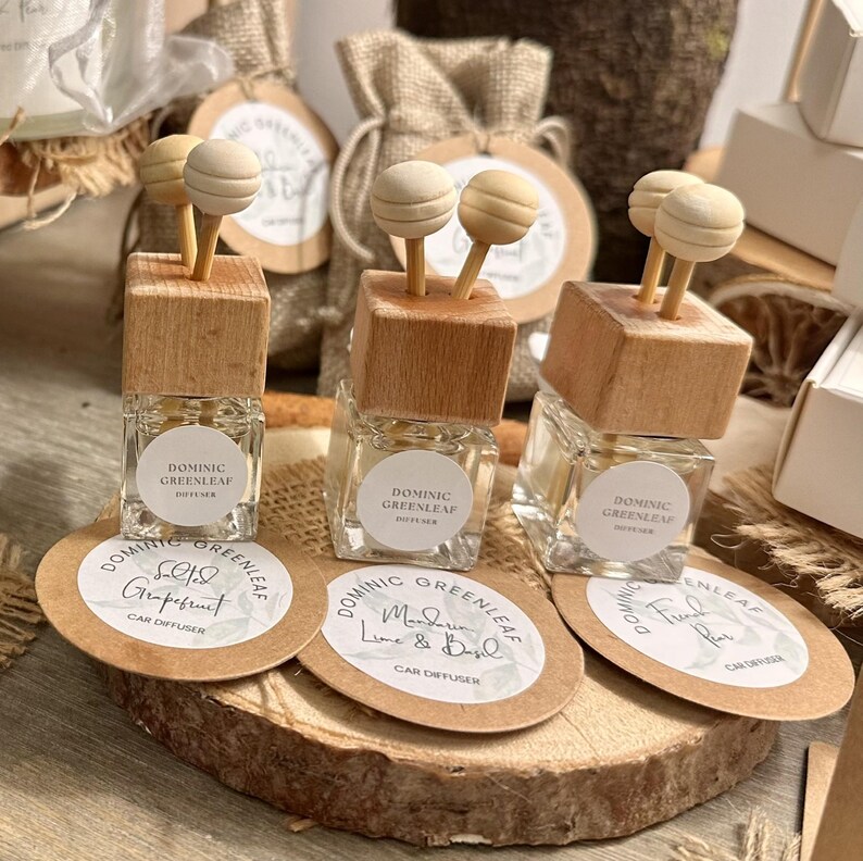 May include: Three small glass car diffusers with wooden tops and wooden ball sticks. Each diffuser has a label that reads "DOMINIC GREENLEAF" and a scent name. The diffusers sit on a wooden slice with round labels.