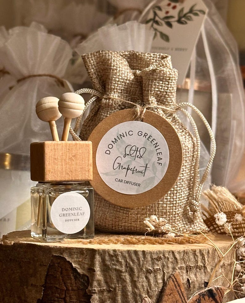 May include: A clear glass car diffuser with a wooden top and two wooden sticks, labeled "Dominic Greenleaf Diffuser." A burlap bag with a round tag that reads "Dominic Greenleaf Salted Grapefruit Car Diffuser" is also present. The items are displayed on a wooden surface.