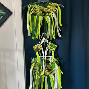 Puede incluir: Conjunto de disfraz de dinosaurio de dos piezas. La parte superior e inferior están hechas de tela de camuflaje con siluetas de dinosaurios. Cintas verdes, blancas y amarillas cuelgan de la parte superior e inferior. El conjunto está colgado en una percha blanca.