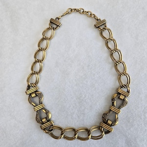May include: A gold-toned metal chain necklace featuring a series of interlocking oval links. The design includes two symmetrical decorative segments with a textured, dotted surface finish, connected by smooth, polished horizontal bars.
