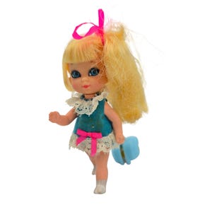 Könnte beinhalten: Eine kleine Puppe mit blondem Haar, das mit einem leuchtend rosa Band zusammengebunden ist, blauen Augen und einem blaugrünen Kleid mit weißem Spitzenkragen und Saum. Das Kleid ist mit einer kleinen rosa Schleife an der Taille verziert, helle Haut und weiße Schuhe.