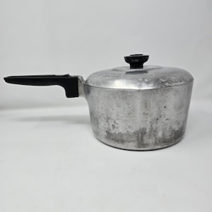 May include: A silver-coloured aluminium saucepan with a matte finish and visible surface wear. The pot features a long, black plastic handle extending from the side and a matching black plastic knob centred on the circular, removable lid.