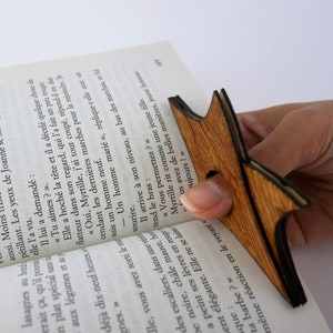 May include: A wooden bookmark shaped like a star with a dark brown stain. The bookmark is being held by a hand over an open book.
