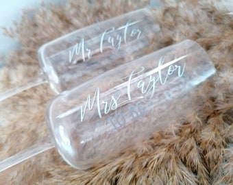 May include: Two clear glass champagne flutes with the text "Mr. Taylor" and "Mrs. Taylor to be" etched on them. The flutes are resting on a bed of dried pampas grass.