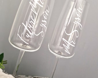 May include: Two clear champagne flutes with white lettering. The flute on the left says "Future Mrs.  [Last Name]" and the flute on the right says "Mr. [Last Name]"