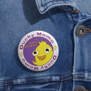 May include: A round button with the text "Ducky Momo" in pink around the edge. The center features a yellow duck character with the text "I HATE PEOPLE!" and a purple background. The button is pinned to a denim jacket.