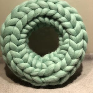 Könnte beinhalten: Ein hellgrüner Strickkranz aus dickem Garn. Der Kranz ist rund und hat eine geflochtene Textur.