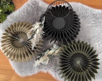 Könnte beinhalten: Drei kreisförmige Wabenpapier-Dekorationen in Beige, Schwarz und Olivgrün, begleitet von einer schwarzen Drahtkäfig-Laterne. Die schwarze Laterne ist mit einem kleinen Arrangement aus getrockneten weißen Blumen verziert.
