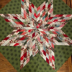 Może przedstawiać: Ręcznie robiona świąteczna kołdra w kształcie gwiazdy z zielonym tłem i dużą gwiazdą pośrodku. Gwiazda wykonana jest z różnych wzorzystych tkanin w kolorach czerwonym, białym i zielonym. Tło ma powtarzający się wzór płatka śniegu.