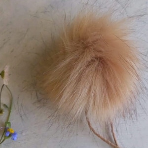Może przedstawiać: Puszysty, jasnobrązowy pompon z imitacji futra o naturalnym wyglądzie. Pompon ma gęstą fakturę i jest przymocowany do cienkiego, jasnobrązowego sznurka. Tło jest jasne i neutralne.