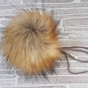 Może przedstawiać: Puszysty brązowo-czarny pompon z imitacji futra z przymocowaną brązową sznurówką.