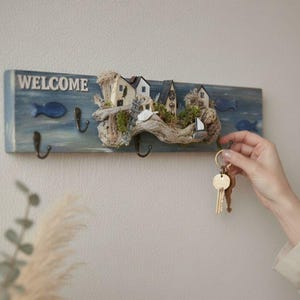 Può includere: Un portachiavi in legno blu con la scritta "WELCOME" in bianco. Il portachiavi presenta case in miniatura, una barca a vela e disegni di pesci. Tre ganci neri sono attaccati per le chiavi.
