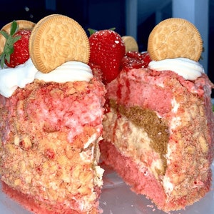 May include: A strawberry shortcake with a slice cut out, revealing layers of pink cake, cream filling, and a crumbly topping. The cake is decorated with fresh strawberries and Oreo cookies.