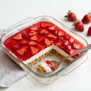 以下が含まれることがあります： イチゴチーズケーキが入った正方形のガラス製のベーキングディッシュ。デザートは、グラハムクラッカーの皮、クリーミーな白いフィリング、赤いグレーズでスライスされたイチゴのトッピングが特徴です。スプーンがチーズケーキの一部をすくいます。新鮮なイチゴが皿の周りに散らばっています。