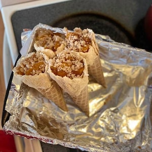 May include: Four waffle cones filled with a peach cobbler mixture, topped with a crumbly topping, and arranged in a foil-lined baking dish. The dessert has a warm, inviting appearance, suggesting a homemade treat.