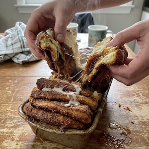 May include: A pull-apart cinnamon bread loaf in a rectangular metal baking pan. The golden-brown pastry is coated in cinnamon sugar, drizzled with white icing, and steaming. The bread is being pulled apart to reveal a soft, layered interior.