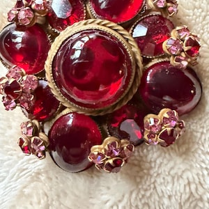 May include: A circular brooch featuring a large central red cabochon stone surrounded by a twisted gold-tone metal border. The piece is accented with smaller red cabochons and clusters of tiny pink faceted rhinestones set in gold-tone metal.