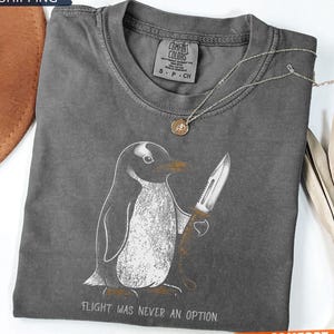 May include: A dark grey short-sleeve t-shirt featuring a graphic of a penguin holding a knife. The text below the penguin reads, "FLIGHT WAS NEVER AN OPTION." The shirt has a crew neckline and a Comfort Colors brand label printed on the inside neck.