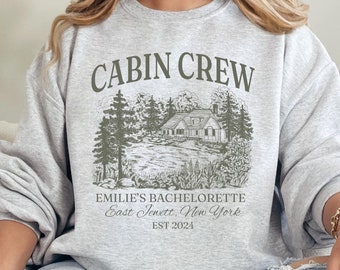 May include: Gray sweatshirt with the text "CABIN CREW" in black lettering. The sweatshirt features a cabin in the woods with trees and a lake. The text "EMILIE'S BACHELORETTE East Jewett, New York EST 2024" is printed below the image.