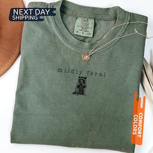 May include: A moss green t-shirt with the words "mildly feral" above a raccoon graphic. The shirt has a crew neck and a small tag on the collar. A gold necklace with a pendant is draped across the shirt. The shirt is folded on a white surface.