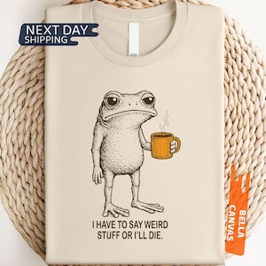 May include: Beige t-shirt featuring a quirky frog illustration holding a mug of coffee. The frog has a grumpy expression. The text "I HAVE TO SAY WEIRD STUFF OR I'LL DIE" is printed below the frog. The shirt is made of natural-colored fabric.