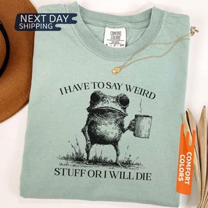 May include: A light green t-shirt with the text "I HAVE TO SAY WEIRD STUFF OR I WILL DIE" and a black illustration of a frog holding a mug. The shirt has a crew neck and short sleeves. The brand tag "COMFORT COLORS" is visible.