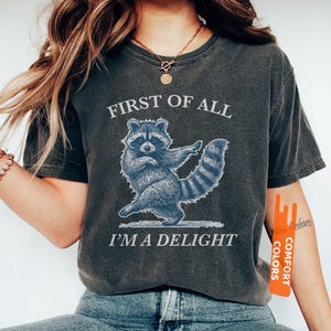 Puede incluir: Camiseta gris oscuro con una ilustraci&oacute;n azul de un mapache y el texto "FIRST OF ALL I'M A DELIGHT". El mapache se representa en una pose de baile. La camiseta est&aacute; hecha de un material suave y c&oacute;modo.