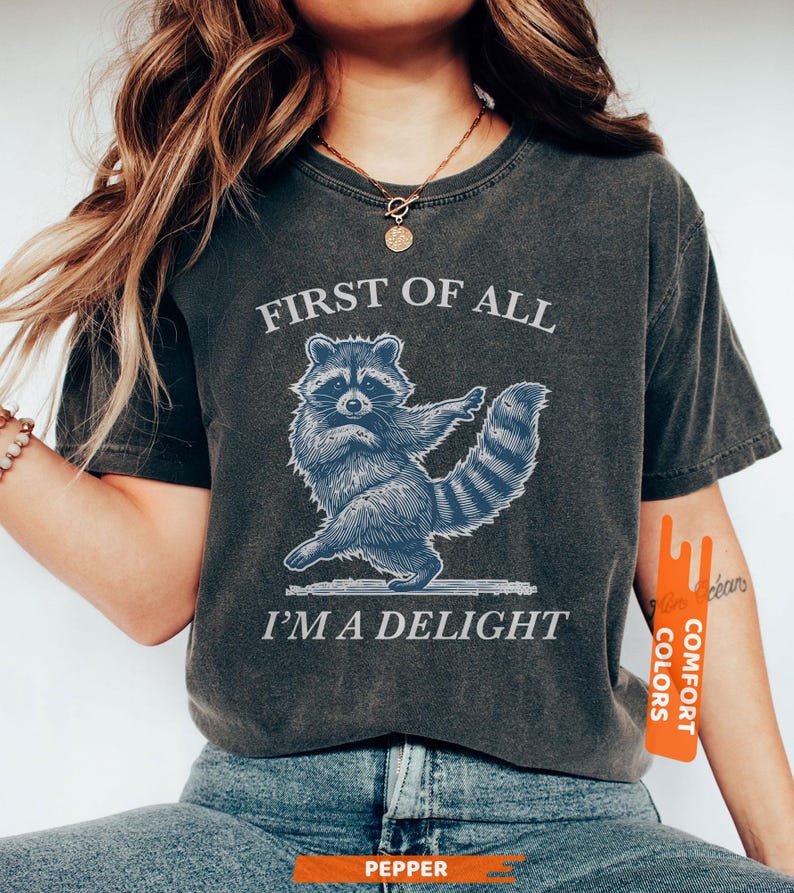 Puede incluir: Camiseta gris oscuro con una ilustraci&oacute;n azul de un mapache y el texto "FIRST OF ALL I'M A DELIGHT". El mapache se representa en una pose de baile. La camiseta est&aacute; hecha de un material suave y c&oacute;modo.