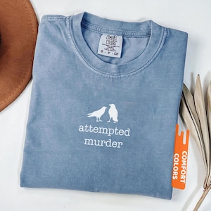 May include: Blue jean coloured t-shirt featuring the words "attempted murder" and a graphic of two birds. The shirt is folded and has a Comfort Colors tag.