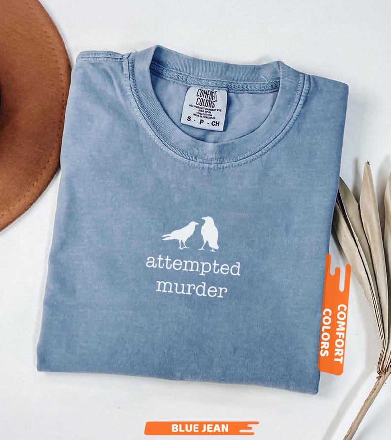 May include: Blue jean coloured t-shirt featuring the words "attempted murder" and a graphic of two birds. The shirt is folded and has a Comfort Colors tag.