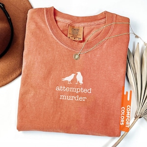 May include: A coral-coloured t-shirt with the words "attempted murder" and a graphic of two birds. The shirt has a crew neck and a small tag that says "Comfort Colors". A gold necklace with a pendant is draped on the shirt.
