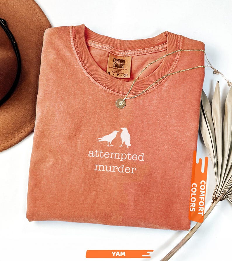 May include: A coral-coloured t-shirt with the words "attempted murder" and a graphic of two birds. The shirt has a crew neck and a small tag that says "Comfort Colors". A gold necklace with a pendant is draped on the shirt.