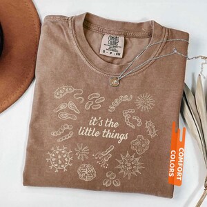 May include: Espresso-colored t-shirt with the phrase "it's the little things" and various microbe illustrations in white. The shirt is from Comfort Colors and has a crew neck. A silver necklace with a small pendant is draped on the shirt.