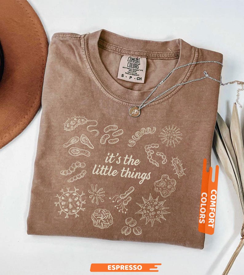 May include: Espresso-colored t-shirt with the phrase "it's the little things" and various microbe illustrations in white. The shirt is from Comfort Colors and has a crew neck. A silver necklace with a small pendant is draped on the shirt.