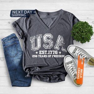 Peut inclure: T-shirt col en V gris fonc&eacute; avec le mot "USA" en lettres blanches us&eacute;es, ainsi que "EST. 1776" et "250 YEARS OF FREEDOM". Le t-shirt est associ&eacute; &agrave; un jean bleu et des baskets grises. Les mots "NEXT DAY SHIPPING" sont dans un encadr&eacute; bleu.