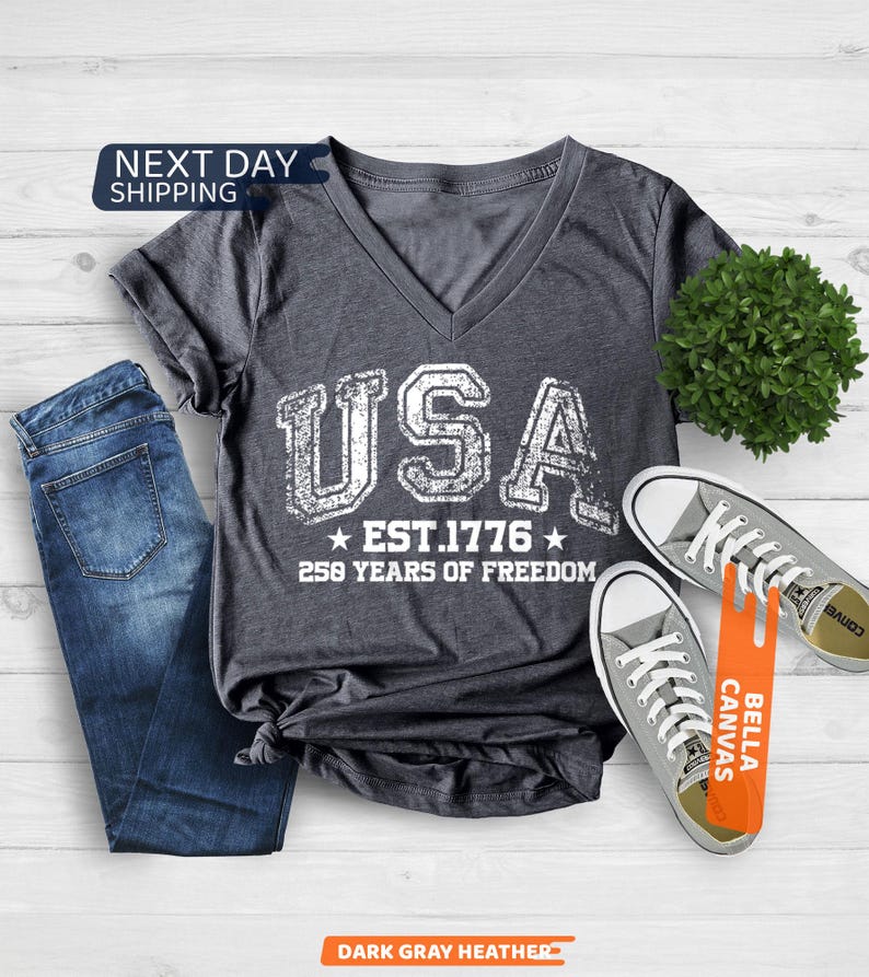 Peut inclure: T-shirt col en V gris fonc&eacute; avec le mot "USA" en lettres blanches us&eacute;es, ainsi que "EST. 1776" et "250 YEARS OF FREEDOM". Le t-shirt est associ&eacute; &agrave; un jean bleu et des baskets grises. Les mots "NEXT DAY SHIPPING" sont dans un encadr&eacute; bleu.