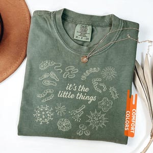 May include: Moss green Comfort Colors t-shirt with the phrase "it's the little things" and various microbe illustrations in white. A gold necklace with a small pendant rests on the shirt.