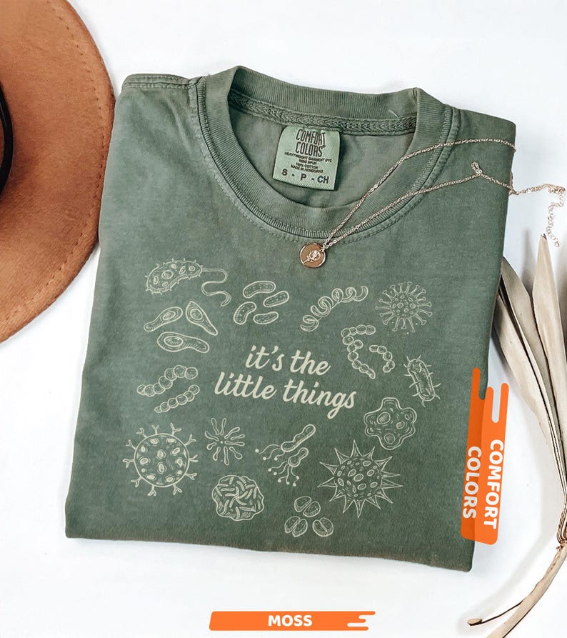 May include: Moss green Comfort Colors t-shirt with the phrase "it's the little things" and various microbe illustrations in white. A gold necklace with a small pendant rests on the shirt.