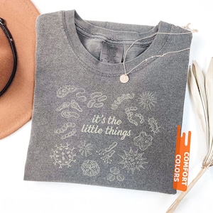 May include: Pepper-colored t-shirt with the phrase "it's the little things" and various microbe illustrations. The shirt has a crew neck and short sleeves. A silver necklace with a small pendant is on the shirt. The shirt is from Comfort Colors.