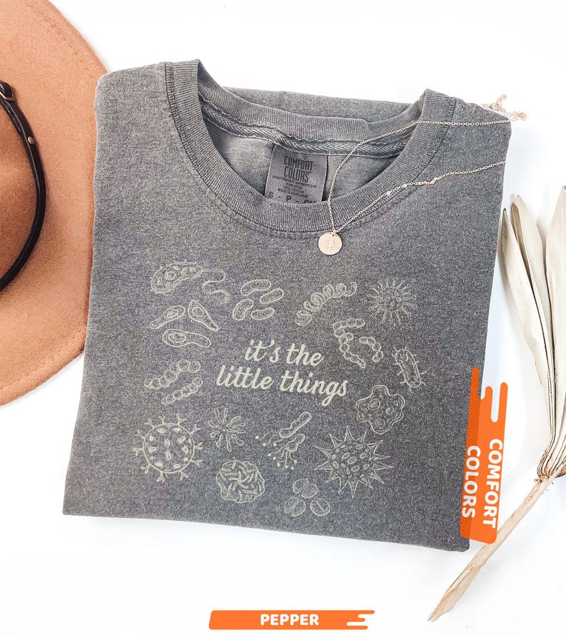 May include: Pepper-colored t-shirt with the phrase "it's the little things" and various microbe illustrations. The shirt has a crew neck and short sleeves. A silver necklace with a small pendant is on the shirt. The shirt is from Comfort Colors.