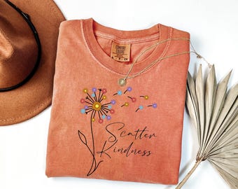 May include: A terracotta-colored short-sleeve t-shirt featuring a graphic of a dandelion with colorful flower heads blowing in the wind. The text "Scatter Kindness" is printed in black script below the flower. The shirt has a crew neckline.