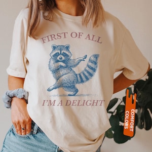 Puede incluir: Camiseta color crema con una ilustraci&oacute;n azul de un mapache bailando. El texto "FIRST OF ALL I'M A DELIGHT" est&aacute; impreso encima y debajo del mapache. La camiseta tiene mangas cortas y cuello redondo.