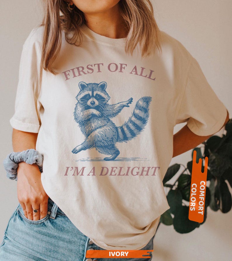 Puede incluir: Camiseta color crema con una ilustraci&oacute;n azul de un mapache bailando. El texto "FIRST OF ALL I'M A DELIGHT" est&aacute; impreso encima y debajo del mapache. La camiseta tiene mangas cortas y cuello redondo.