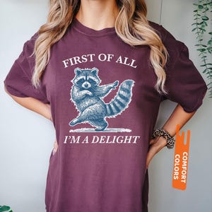 Puede incluir: Camiseta color bayas con un dise&ntilde;o de mapache azul y blanco. El texto "FIRST OF ALL I'M A DELIGHT" es blanco. La camiseta es de la marca Comfort Colors.