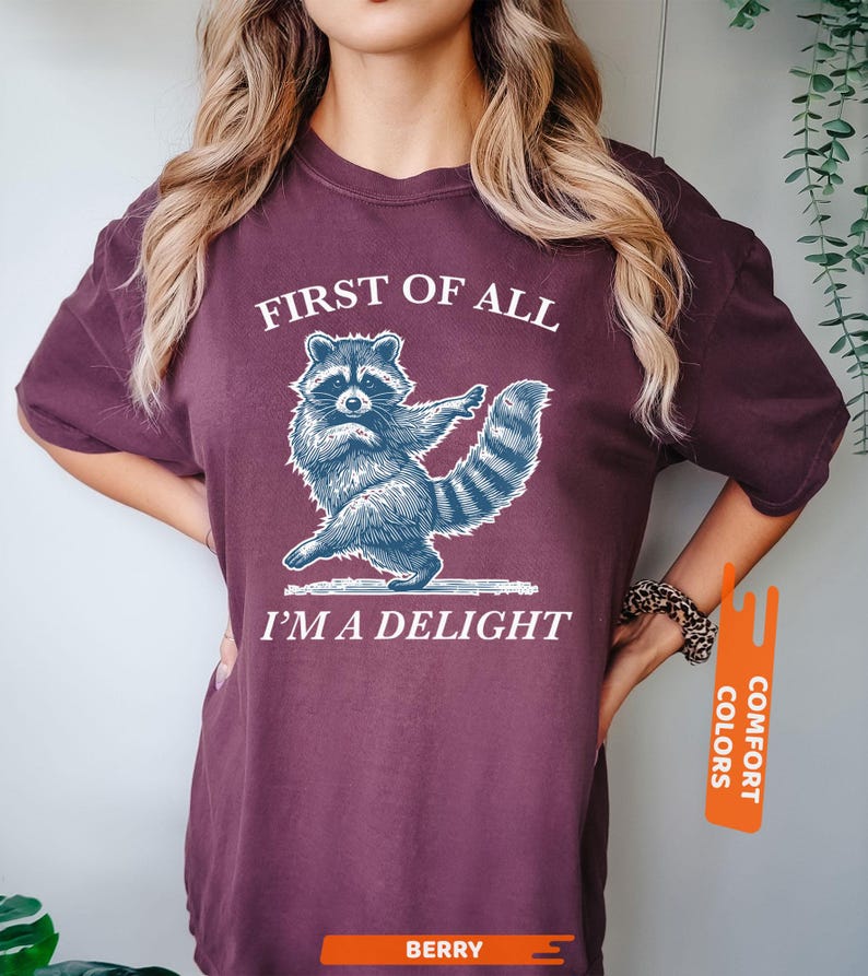 Puede incluir: Camiseta color bayas con un dise&ntilde;o de mapache azul y blanco. El texto "FIRST OF ALL I'M A DELIGHT" es blanco. La camiseta es de la marca Comfort Colors.
