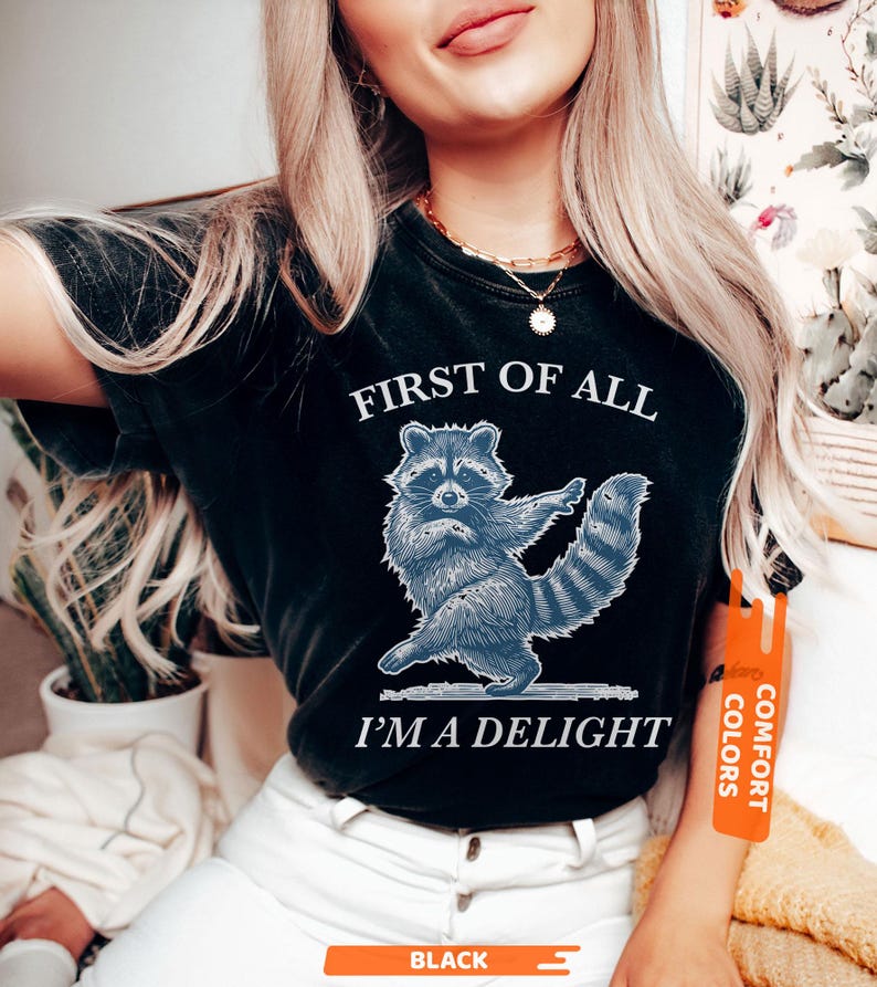 Puede incluir: Camiseta negra con un dise&ntilde;o de mapache azul y el texto "FIRST OF ALL I'M A DELIGHT". La camiseta tiene un aspecto desgastado y una etiqueta Comfort Colors.