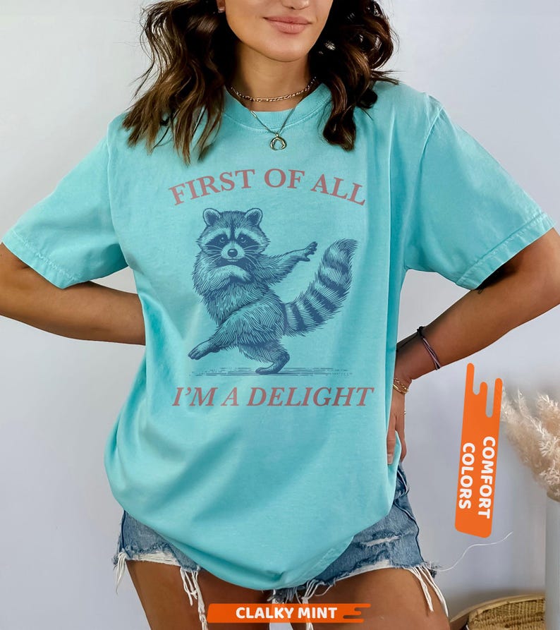 Puede incluir: Camiseta turquesa con el texto "FIRST OF ALL I'M A DELIGHT" y una ilustraci&oacute;n de un mapache bailando. La camiseta tiene mangas cortas y cuello redondo.